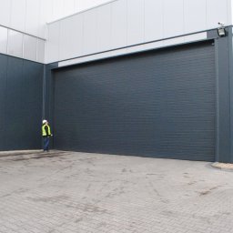 SOLID OKNA - Duża, szara brama przemysłowa z poziomymi panelami, zamontowana w hali. Obok stoi osoba w kasku i kamizelce odblaskowej. Kostka brukowa na podłożu.