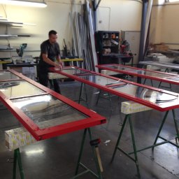 SOLID OKNA - Warsztatowa sceneria: mężczyzna montuje uszczelki w czerwonych ramach okiennych na kozłach. W tle narzędzia i materiały, tworzące autentyczny klimat pracy.