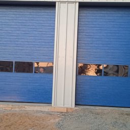 SOLID OKNA - Niebieska brama segmentowa z oknami, oddzielona pionowym, srebrnym profilem. Widoczne elementy konstrukcji budynku i fragment otoczenia.