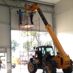 SOLID OKNA - Montaż bramy przemysłowej w hali produkcyjnej. Pracownicy na rusztowaniu i podnośniku teleskopowym. Widoczne elementy konstrukcyjne bramy i narzędzia.