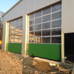 SOLID OKNA - Nowoczesna brama przemysłowa segmentowa z przeszkleniami i zielonym panelem dolnym, zamontowana w hali o srebrnej elewacji. Na pierwszym planie gruz.