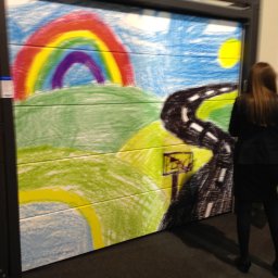 SOLID OKNA - Brama garażowa z rysunkiem dziecka: tęcza, droga i słońce. Kobieta stoi tyłem, obok stolik z napojem. Nietypowa dekoracja bramy garażowej.