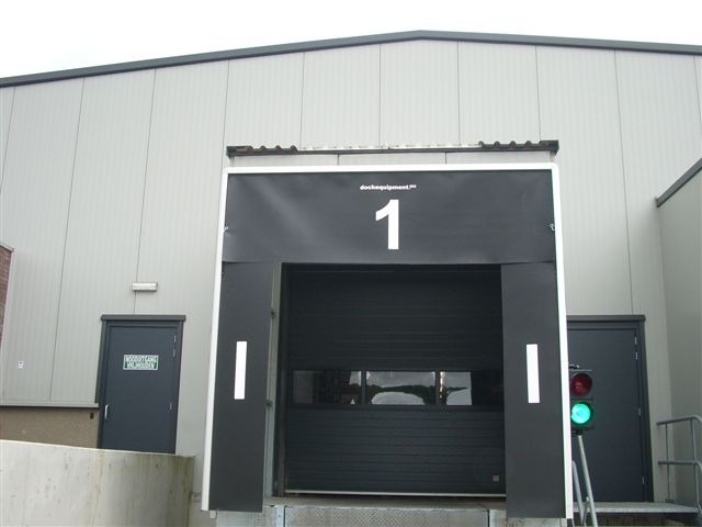 Brama przemysłowa z numerem 1 i sygnalizacją świetlną na tle szarej hali. Widoczny fragment rampy załadunkowej i drzwi wejściowe.