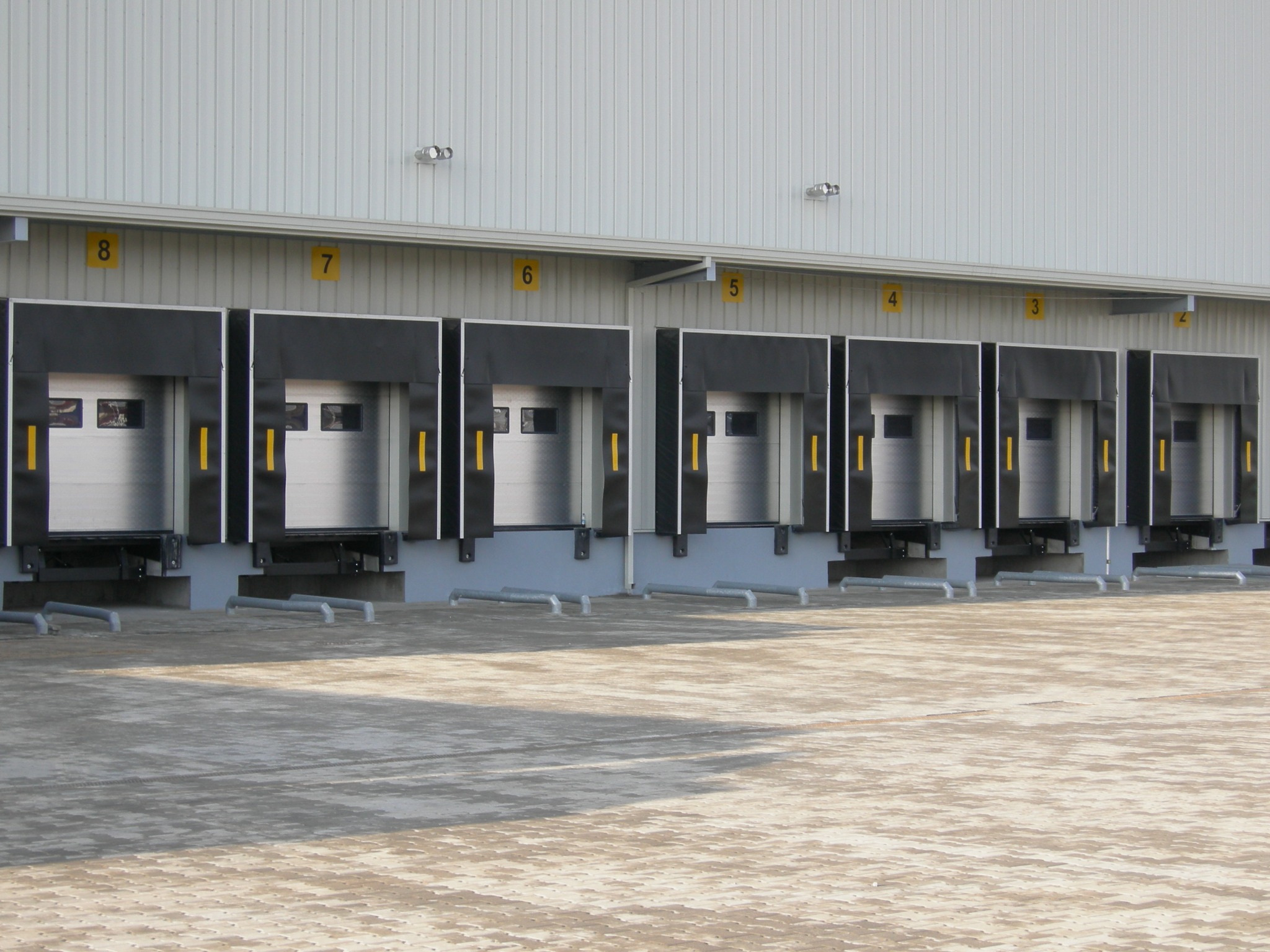 Rząd częściowo otwartych, numerowanych bram przemysłowych z czarnymi uszczelnieniami, widocznych z zewnątrz, na tle białej hali.