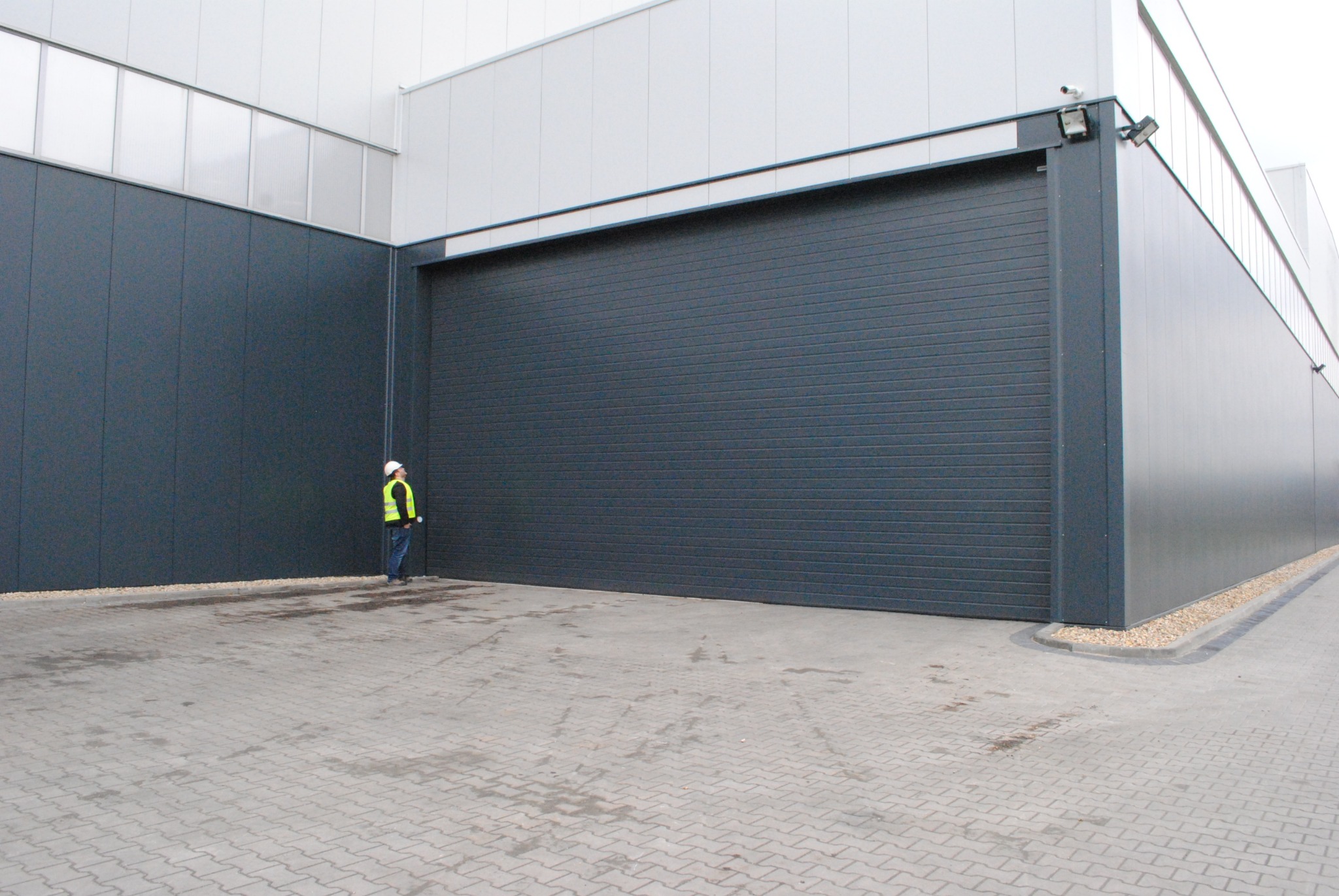Duża, szara brama przemysłowa z poziomymi panelami, zamontowana w hali. Obok stoi osoba w kasku i kamizelce odblaskowej. Kostka brukowa na podłożu.