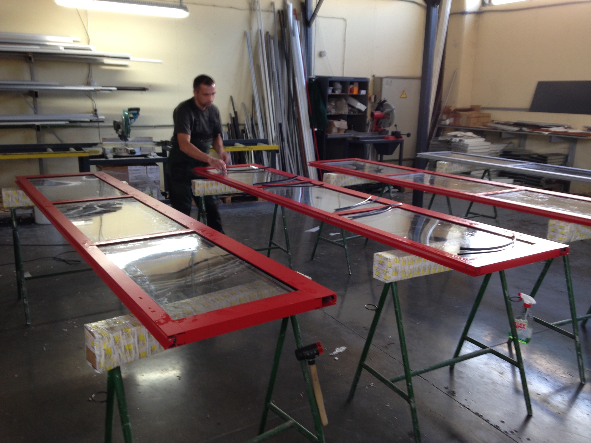 Warsztatowa sceneria: mężczyzna montuje uszczelki w czerwonych ramach okiennych na kozłach. W tle narzędzia i materiały, tworzące autentyczny klimat pracy.