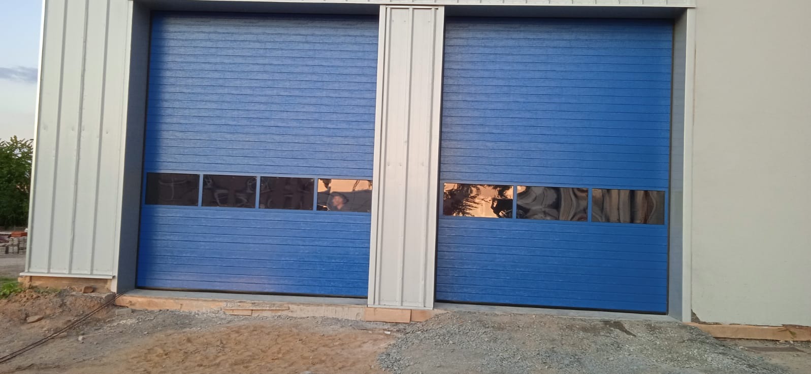 Niebieska brama segmentowa z oknami, oddzielona pionowym, srebrnym profilem. Widoczne elementy konstrukcji budynku i fragment otoczenia.