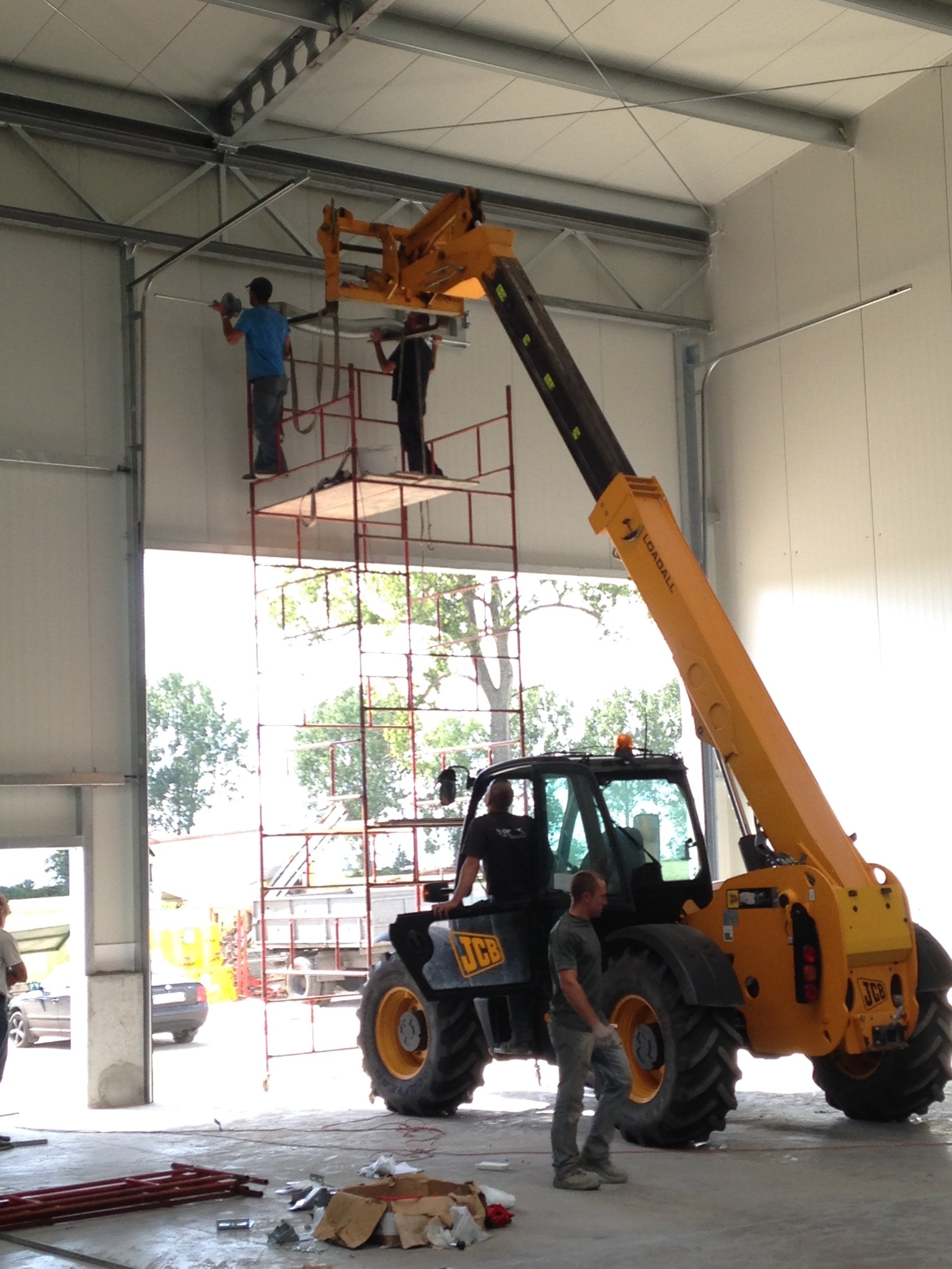 Montaż bramy przemysłowej w hali produkcyjnej. Pracownicy na rusztowaniu i podnośniku teleskopowym. Widoczne elementy konstrukcyjne bramy i narzędzia.