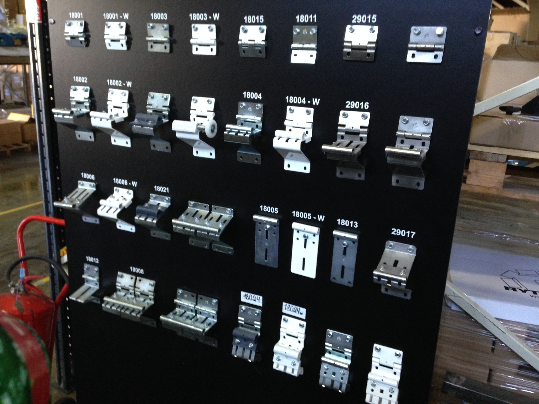 Ekspozycja różnych typów zawiasów do bram garażowych i przemysłowych, zamontowanych na czarnej tablicy z numerami identyfikacyjnymi. Widoczne detale konstrukcyjne.