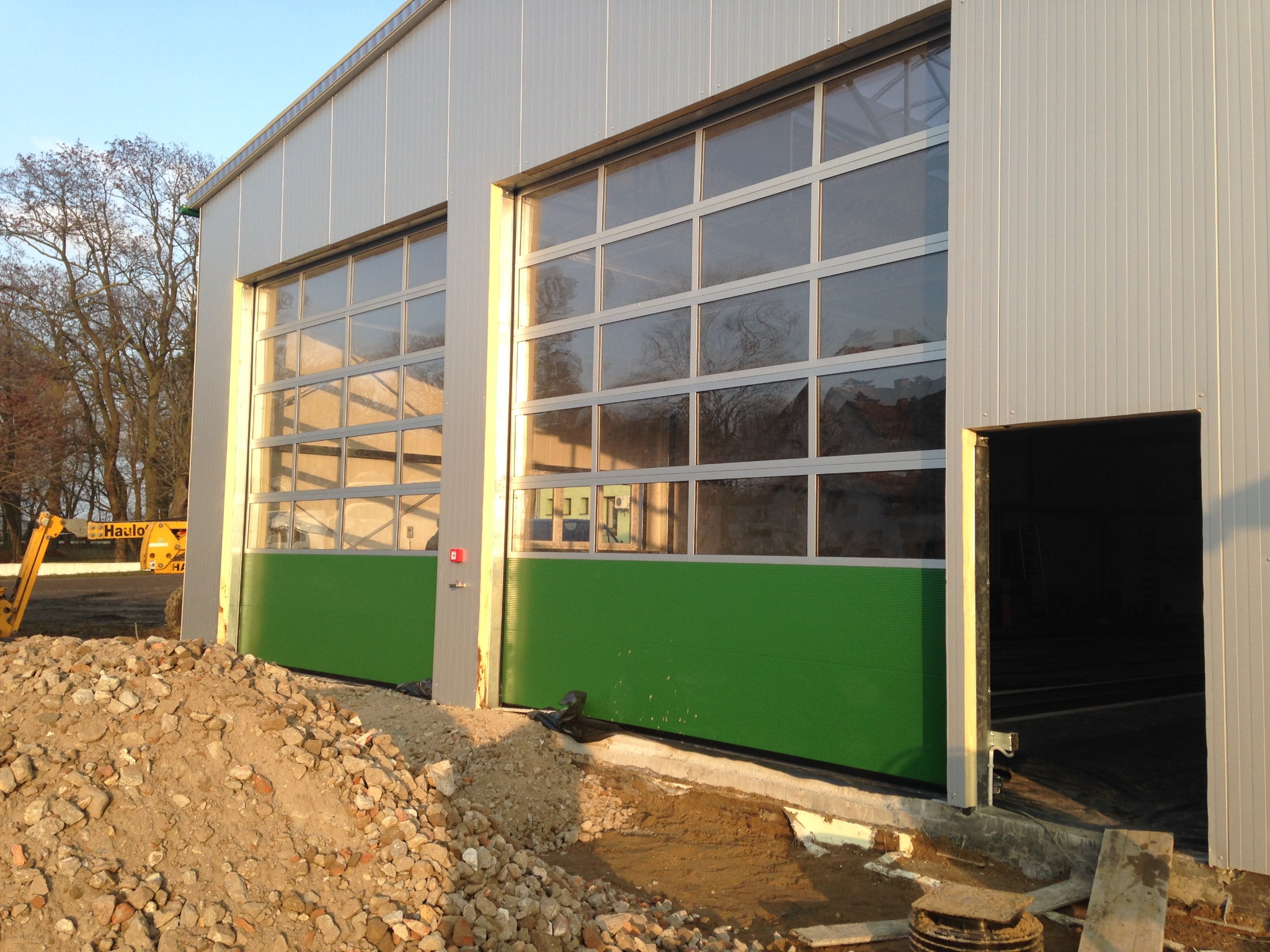 Nowoczesna brama przemysłowa segmentowa z przeszkleniami i zielonym panelem dolnym, zamontowana w hali o srebrnej elewacji. Na pierwszym planie gruz.