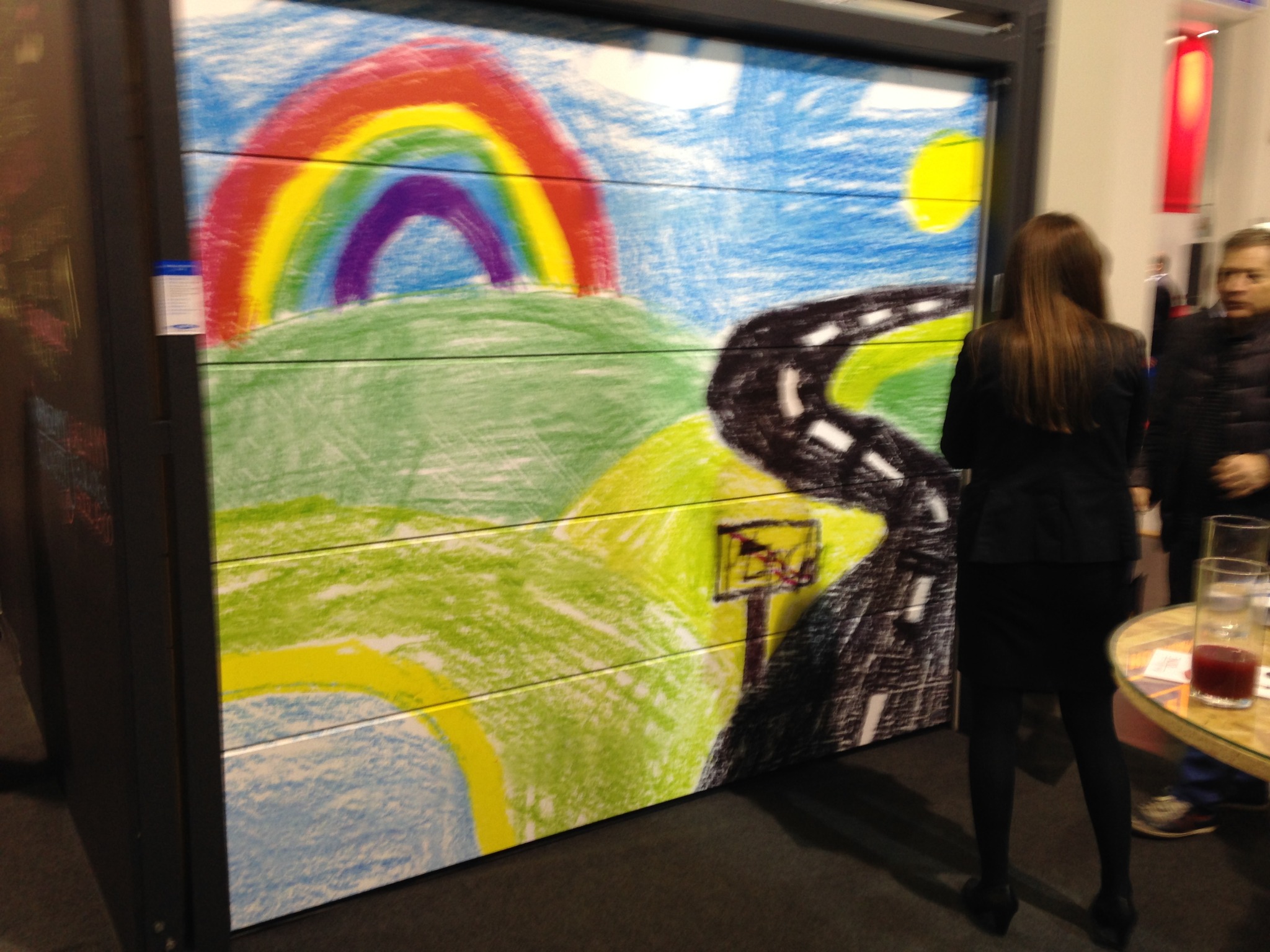 Brama garażowa z rysunkiem dziecka: tęcza, droga i słońce. Kobieta stoi tyłem, obok stolik z napojem. Nietypowa dekoracja bramy garażowej.