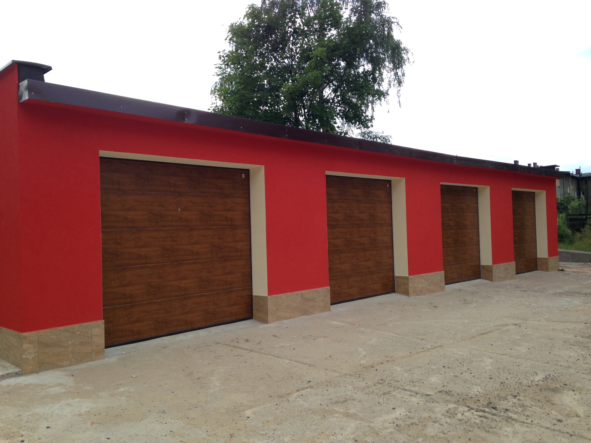 Czerwony budynek garażowy z czterema drewnianymi bramami segmentowymi, wykończony płytkami imitującymi marmur. Widok z zewnątrz, w tle drzewo i budynki.