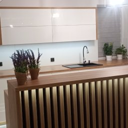 Iskra - Nowoczesna kuchnia z białymi szafkami i drewnianym blatem. Wyspa z oświetleniem LED. Uwagę przykuwają dekoracje i rośliny doniczkowe. Aranżacja pod skosem dachu.