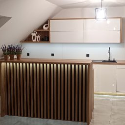 Iskra - Nowoczesna kuchnia na poddaszu z białymi szafkami i drewnianym blatem. Uwagę przykuwa podświetlany, lamelowy barek i dekoracje na półce.