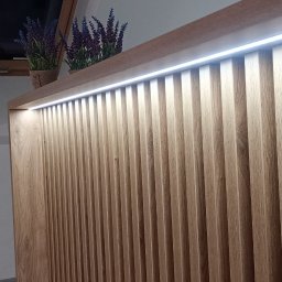 Iskra - Drewniana szafa z lamelami i oświetleniem LED, zwieńczona dekoracyjnymi kwiatami w doniczkach. Ustawiona na poddaszu, widoczne okno dachowe.