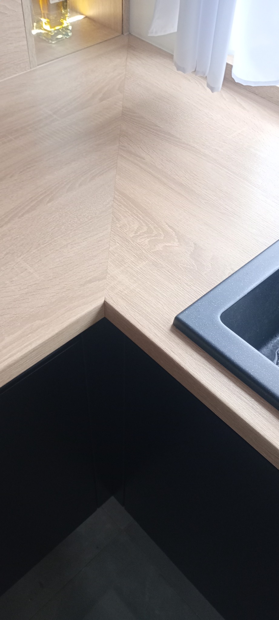 Fragment blatu kuchennego z jasnego drewna i czarnego zlewu, oświetlonego naturalnym światłem. Widoczny narożnik blatu i fragment szafki.