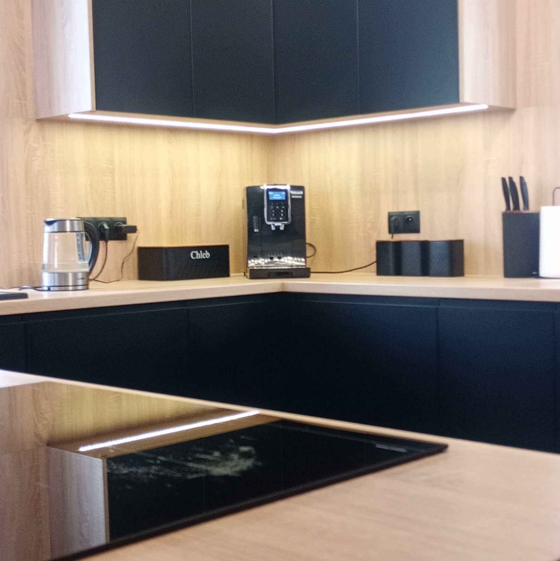 Nowoczesna kuchnia z drewnianymi blatami i czarnymi szafkami. Podświetlenie LED pod szafkami górnymi. Na blacie ekspres do kawy i czajnik.