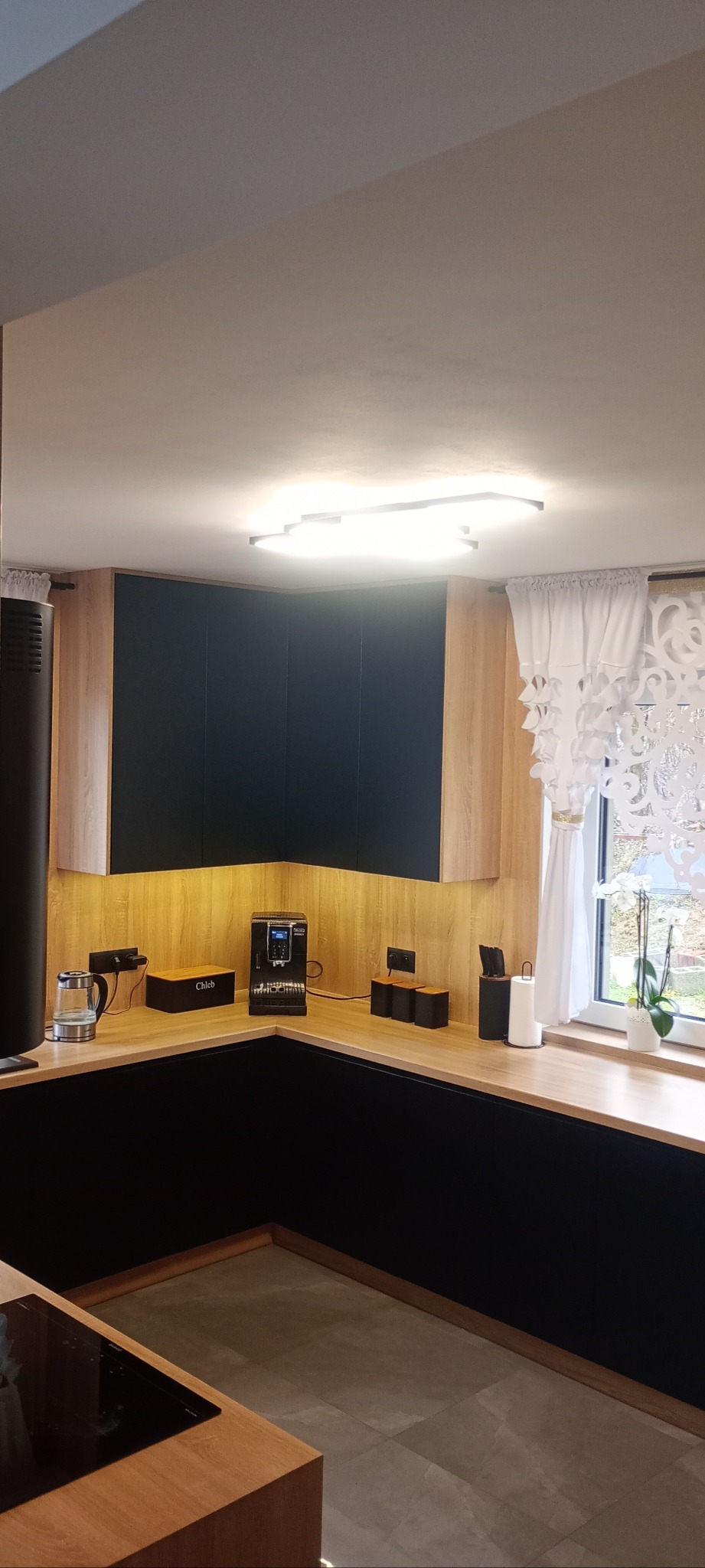 Nowoczesna kuchnia z czarnymi frontami i drewnianymi blatami, oświetlona ciepłym światłem. Widoczny ekspres do kawy i akcesoria kuchenne. Minimalistyczny design.