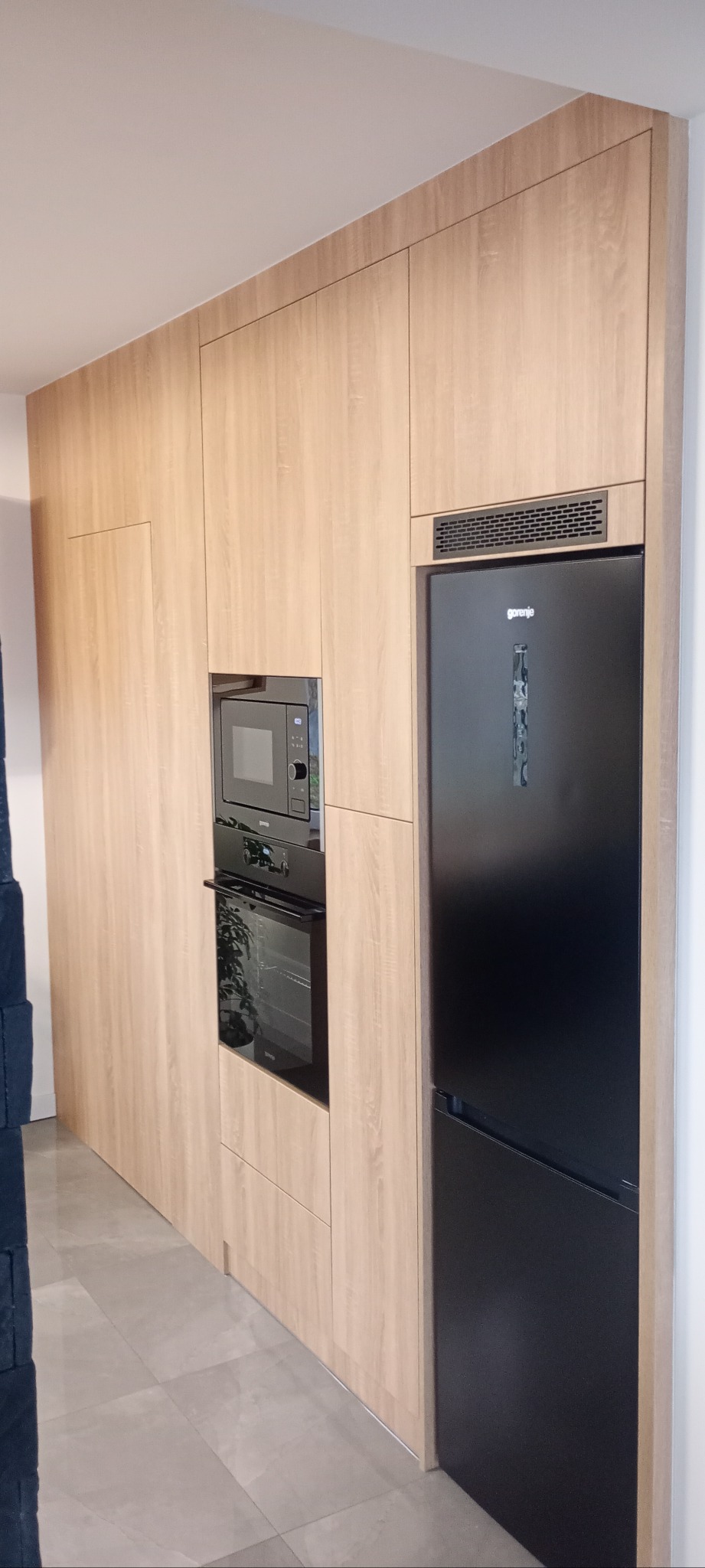 Jasna zabudowa kuchenna z drewnopodobnym frontem, wkomponowana czarna lodówka i piekarnik z mikrofalówką. Minimalistyczny design, szare płytki podłogowe.