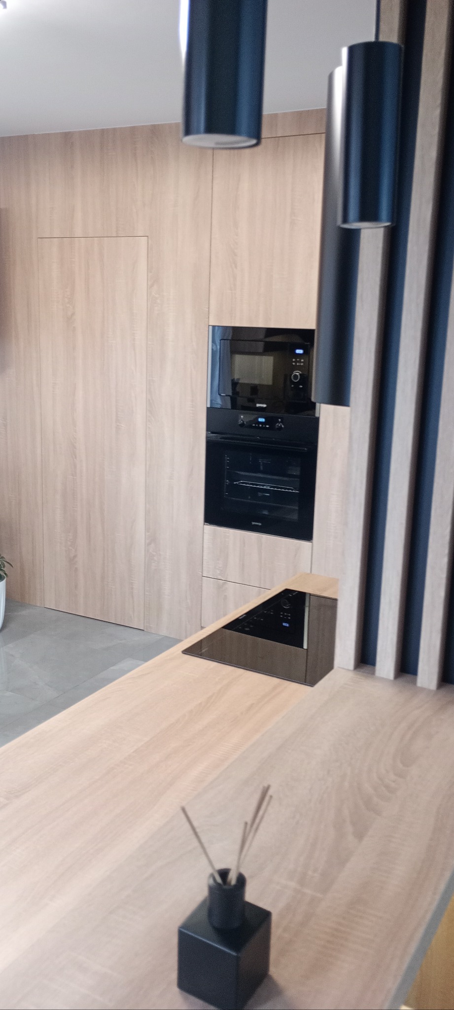 Nowoczesna kuchnia z drewnianymi frontami, czarnym piekarnikiem i płytą indukcyjną. Minimalistyczne lampy wiszące i dekoracyjny dyfuzor zapachowy na blacie.