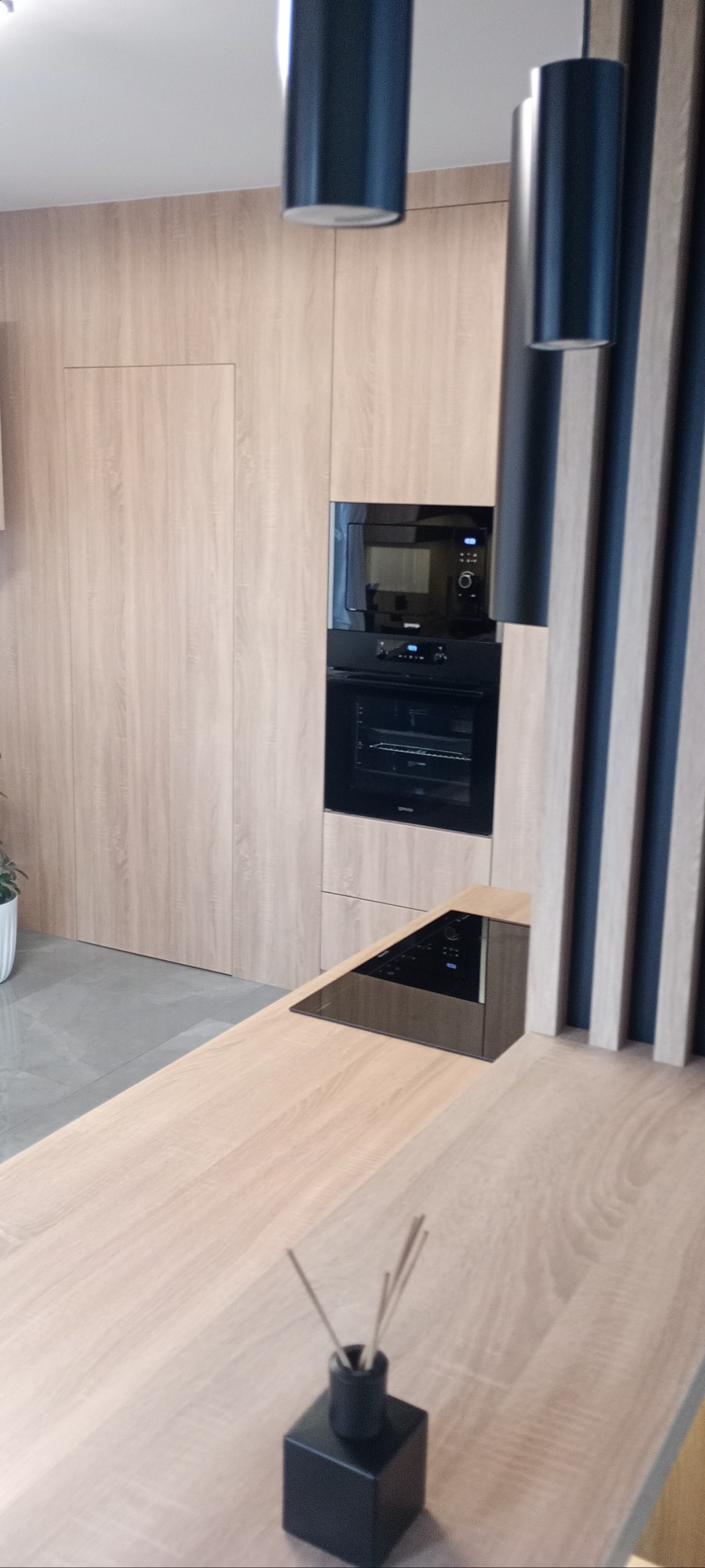 Nowoczesna kuchnia z drewnianymi frontami, czarnym sprzętem AGD i dekoracyjnymi lamelami. Na blacie dyfuzor zapachowy. Minimalistyczny design.