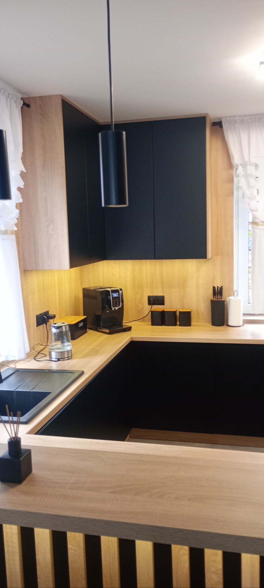 Nowoczesna kuchnia z czarnymi szafkami i drewnianym blatem, oświetlona ciepłym światłem. Widoczny ekspres do kawy i akcesoria kuchenne. Minimalistyczny design.