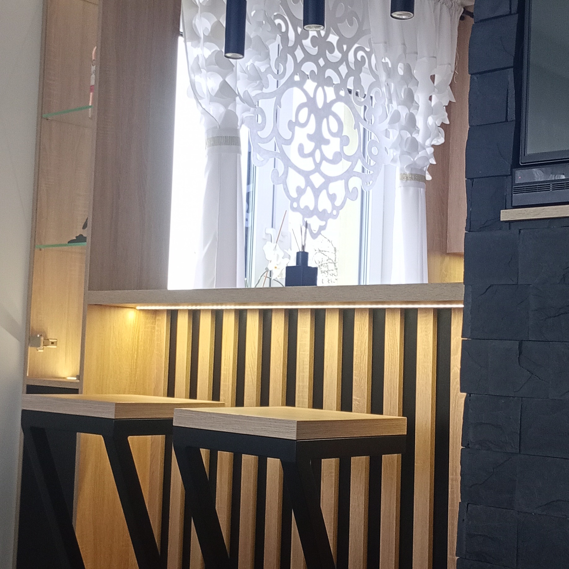 Nowoczesna kuchnia z barkiem: drewniane lamele podświetlone LED, czarne hokery z drewnianymi siedziskami, dekoracyjna zasłona i ściana z czarnej cegły.