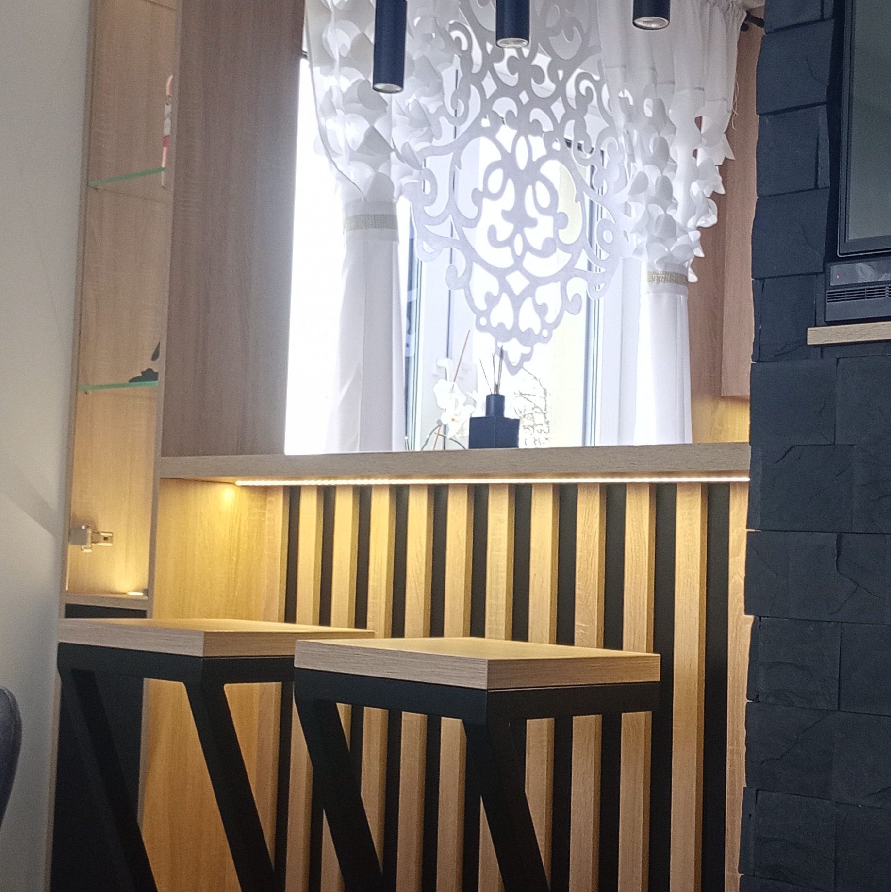 Nowoczesna aranżacja baru w kuchni z drewnianymi hokerami na tle dekoracyjnej ściany z kamienia i ażurowej zasłony. Oświetlenie LED podkreśla detale.