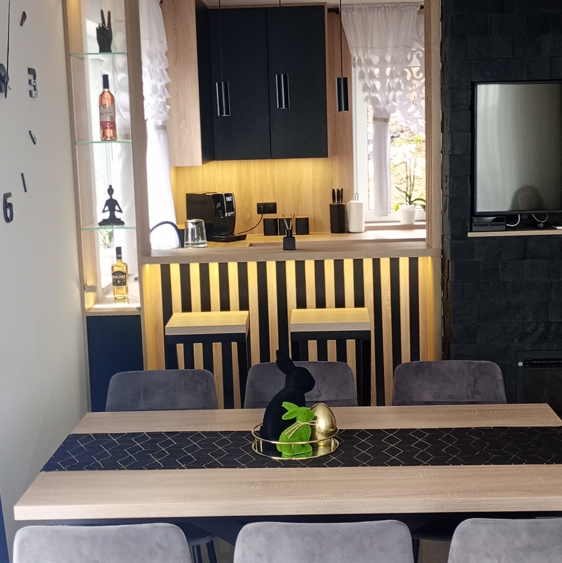 Nowoczesna kuchnia z barkiem i czarnymi szafkami, oświetlona listwą LED. Stół z dekoracją wielkanocną i szarymi krzesłami. Minimalistyczny design.