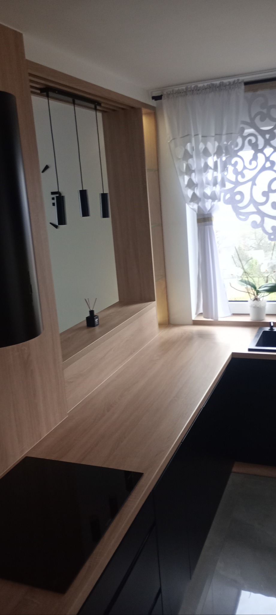 Nowoczesna kuchnia z drewnianym blatem, czarnymi szafkami i minimalistycznym oświetleniem. Widoczny fragment płyty indukcyjnej i dekoracyjny dyfuzor zapachowy.