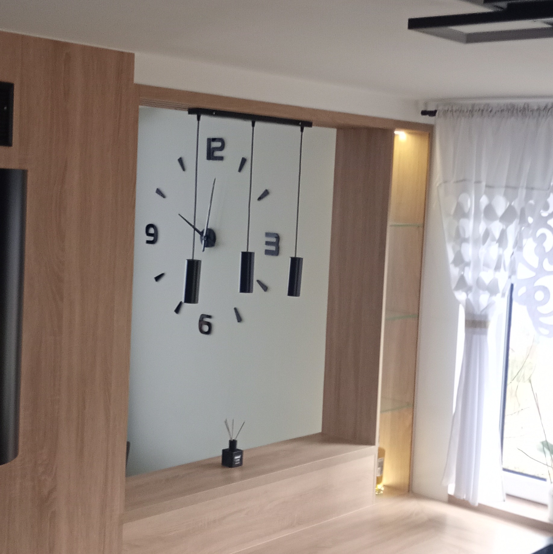 Nowoczesna zabudowa meblowa z drewnopodobnym dekorem, wkomponowana w ścianę. Na ścianie dekoracyjny zegar z wiszącymi elementami. W tle okno z białą firanką.