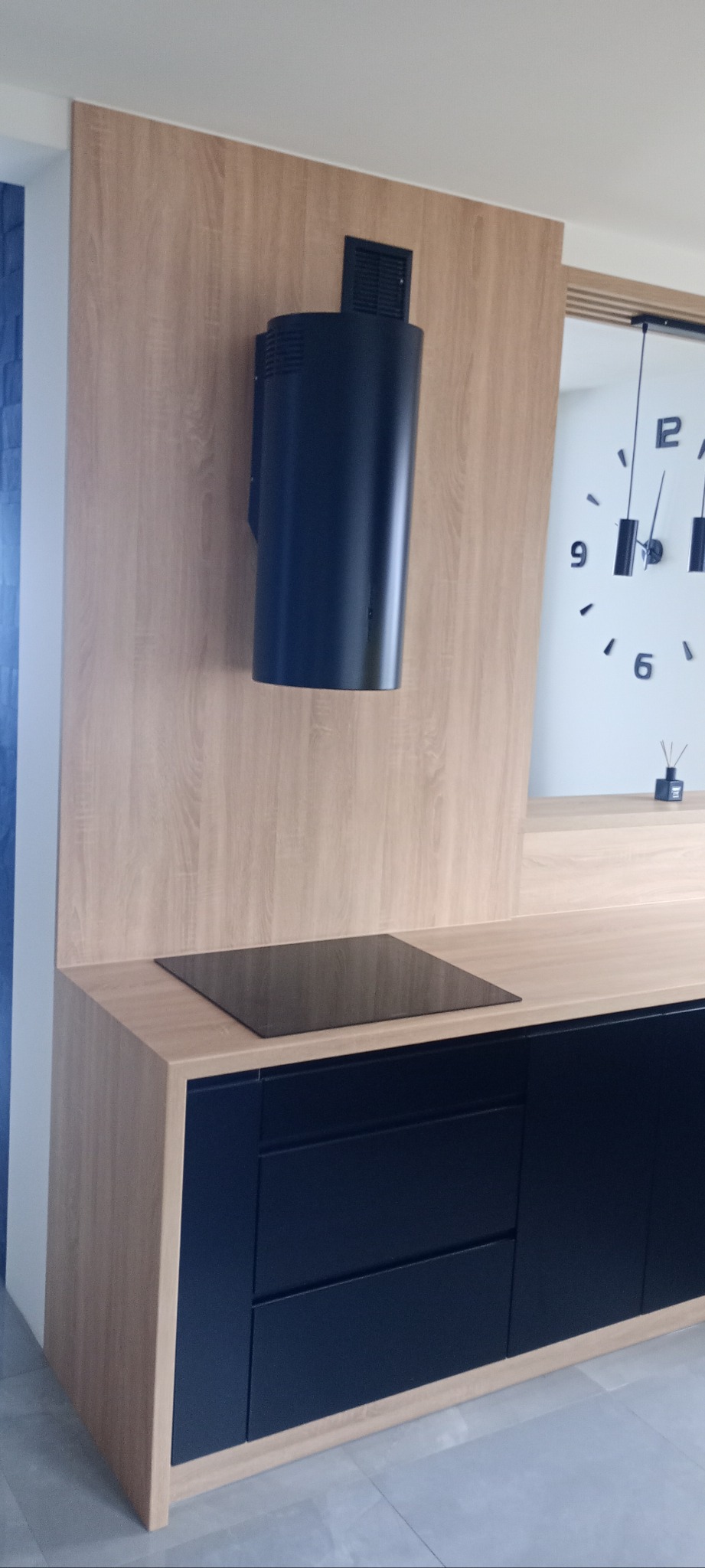 Nowoczesna kuchnia z czarnymi frontami szafek i drewnianym blatem. Na blacie płyta indukcyjna, nad nią czarny okap. W tle zegar na ścianie.