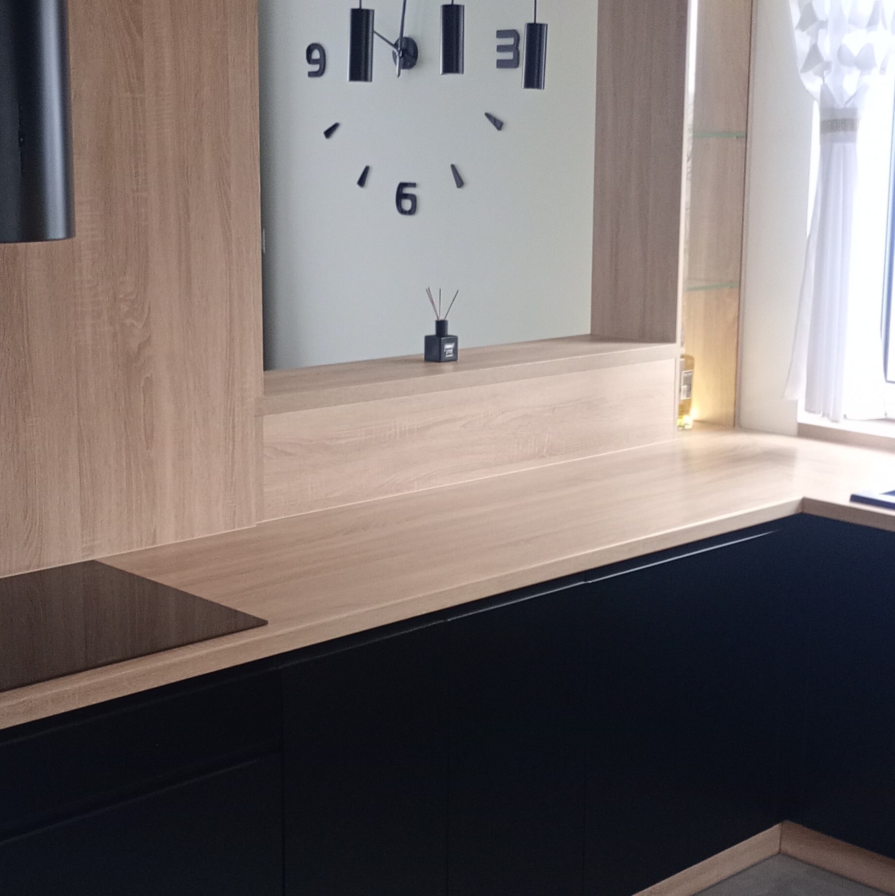 Nowoczesna kuchnia z czarnymi szafkami i drewnianym blatem. Na ścianie duży zegar i dekoracyjny dyfuzor zapachowy. Minimalistyczny design.