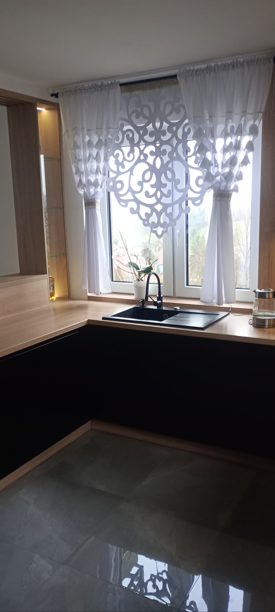 Nowoczesna kuchnia z czarnymi szafkami i drewnianym blatem, okno z dekoracyjną firanką, czarny zlew i bateria. Minimalistyczny design, jasne wnętrze.