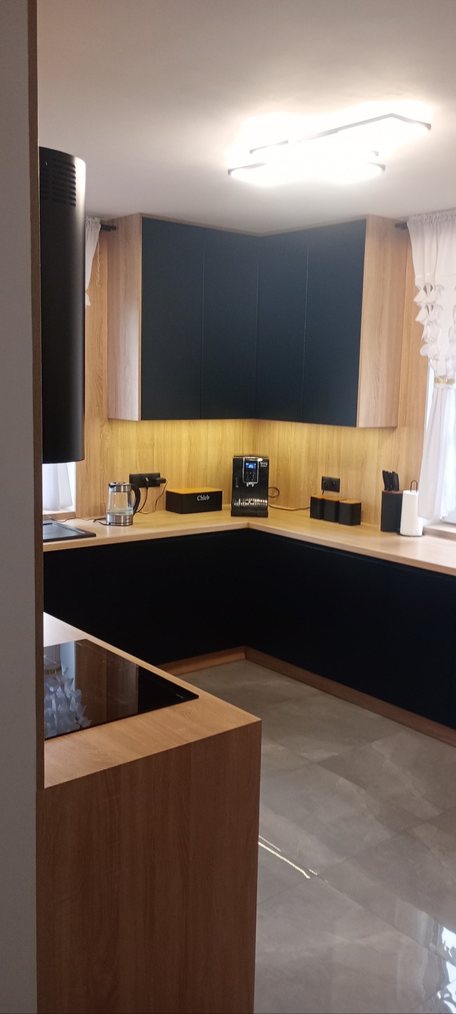 Nowoczesna kuchnia z czarnymi frontami i drewnianymi akcentami, oświetlenie LED pod szafkami, ekspres do kawy na blacie. Minimalistyczny design.