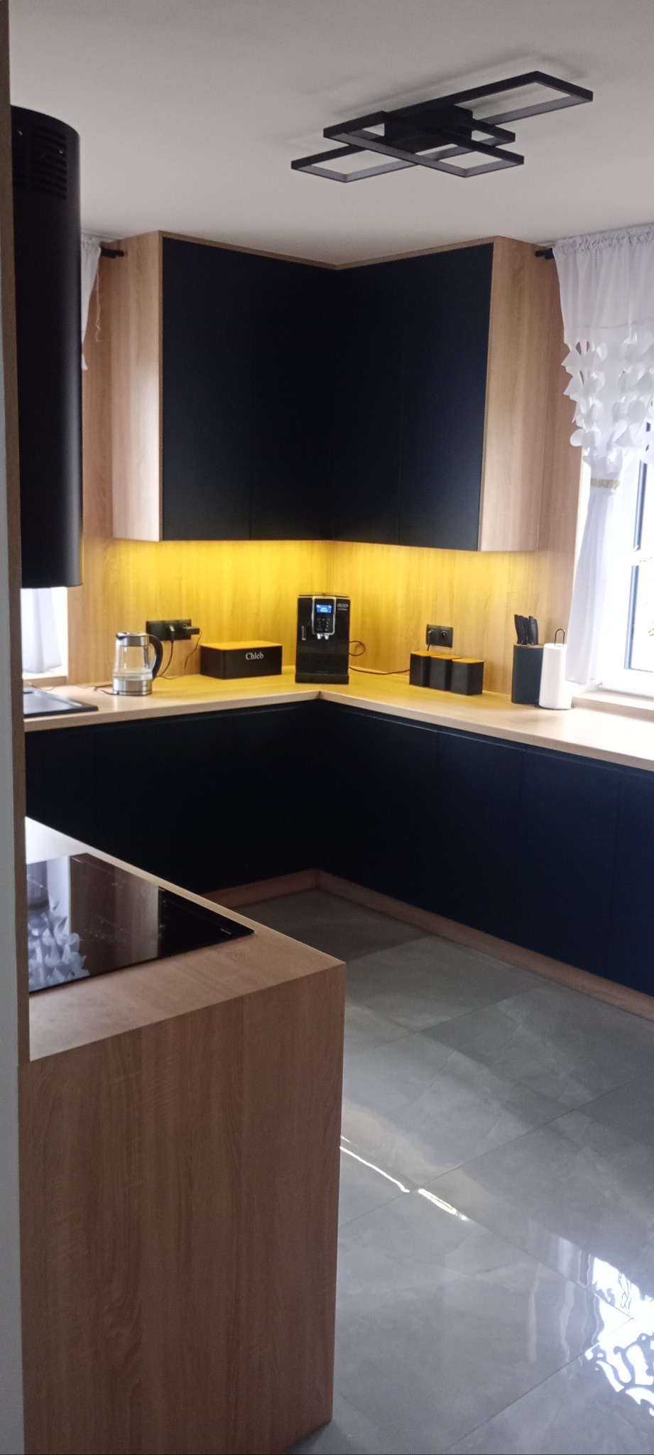 Nowoczesna kuchnia z czarnymi szafkami i drewnianymi akcentami, oświetlona ciepłym światłem LED. Widoczny ekspres do kawy i akcesoria kuchenne.