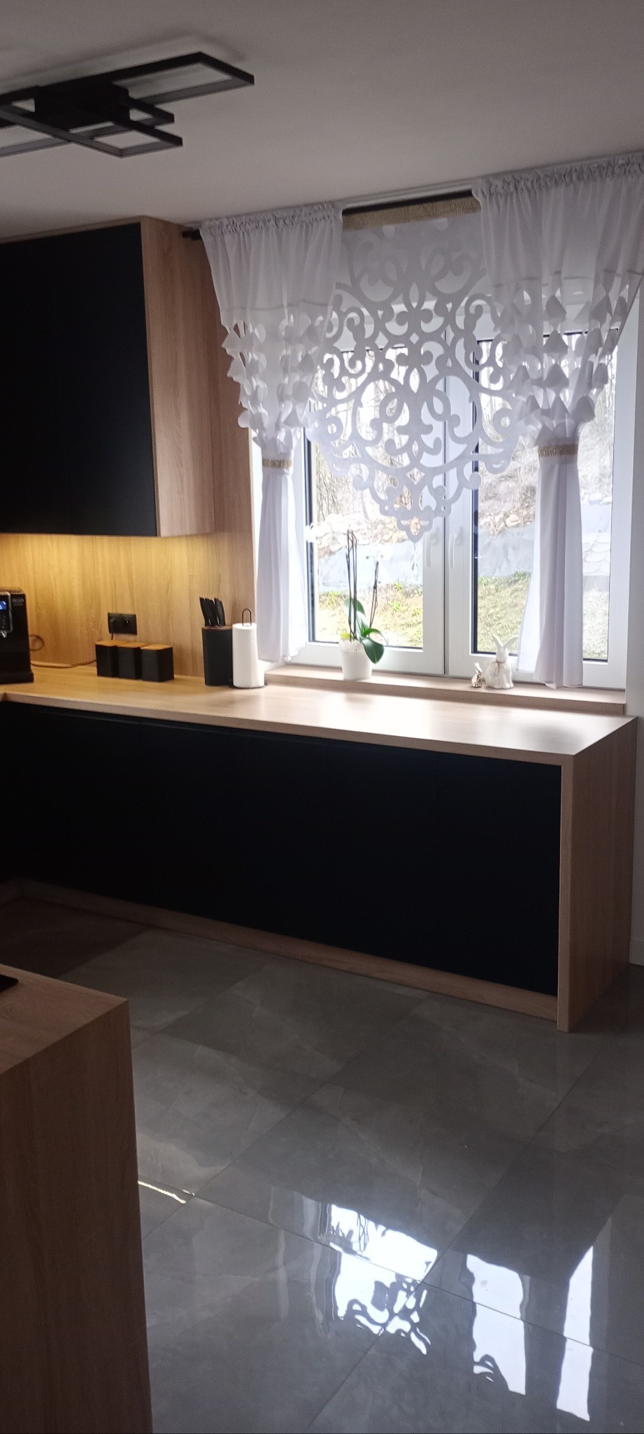 Nowoczesna kuchnia z czarnymi frontami i drewnianym blatem pod oknem z dekoracyjną, białą firanką. Na blacie kwiaty i ozdoby. Szare płytki podłogowe.