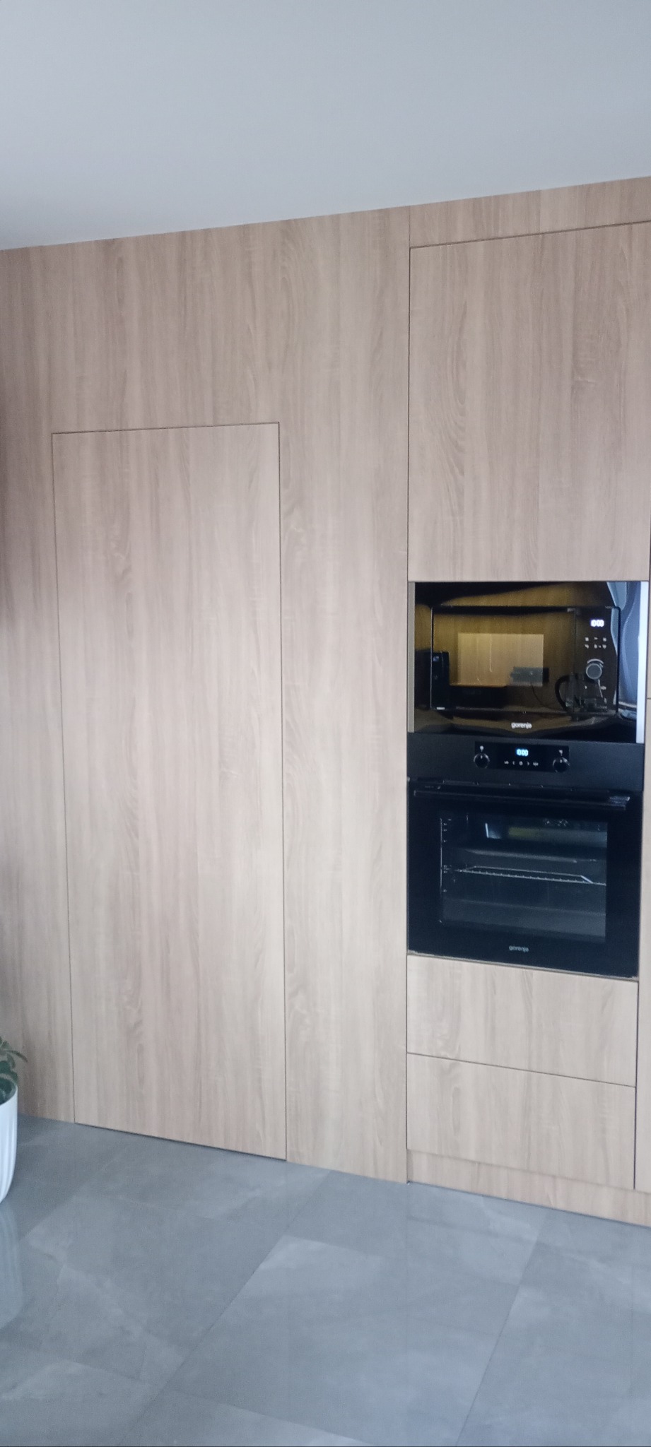 Jasna, drewnopodobna zabudowa kuchenna z ukrytymi drzwiami i wbudowanym piekarnikiem Gorenje. Minimalistyczny design, szare płytki podłogowe.