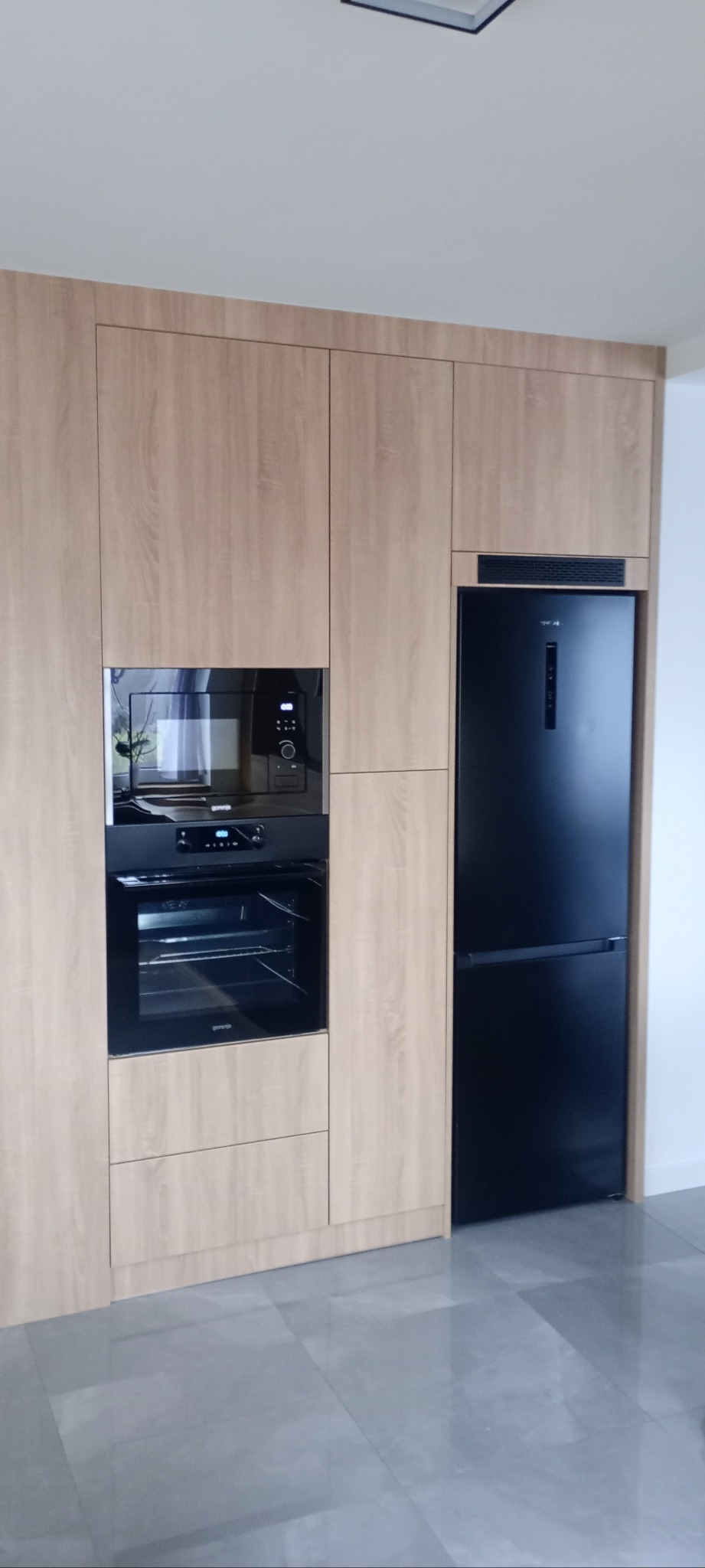 Jasne drewniane szafki kuchenne z czarnym piekarnikiem, mikrofalówką i lodówką. Minimalistyczny design, nowoczesne wykończenie, szare płytki podłogowe.