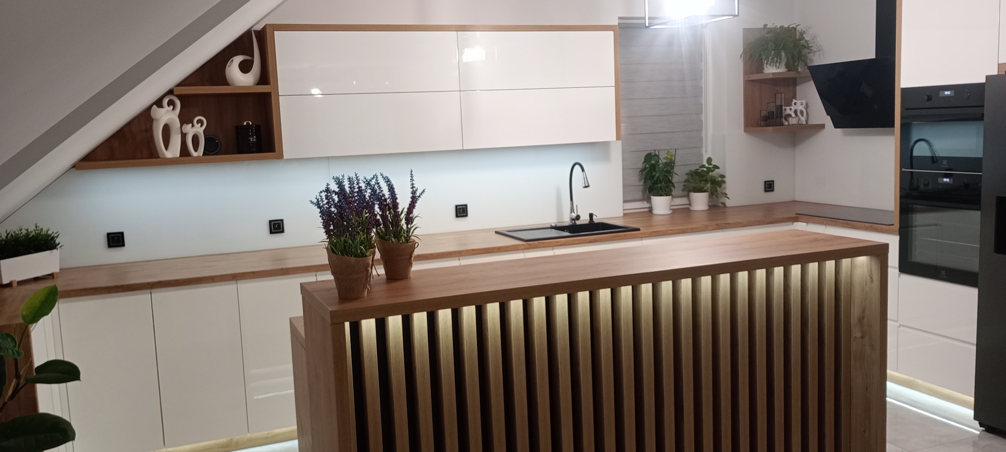 Nowoczesna kuchnia z białymi szafkami i drewnianym blatem. Wyspa z oświetleniem LED. Uwagę przykuwają dekoracje i rośliny doniczkowe. Aranżacja pod skosem dachu.