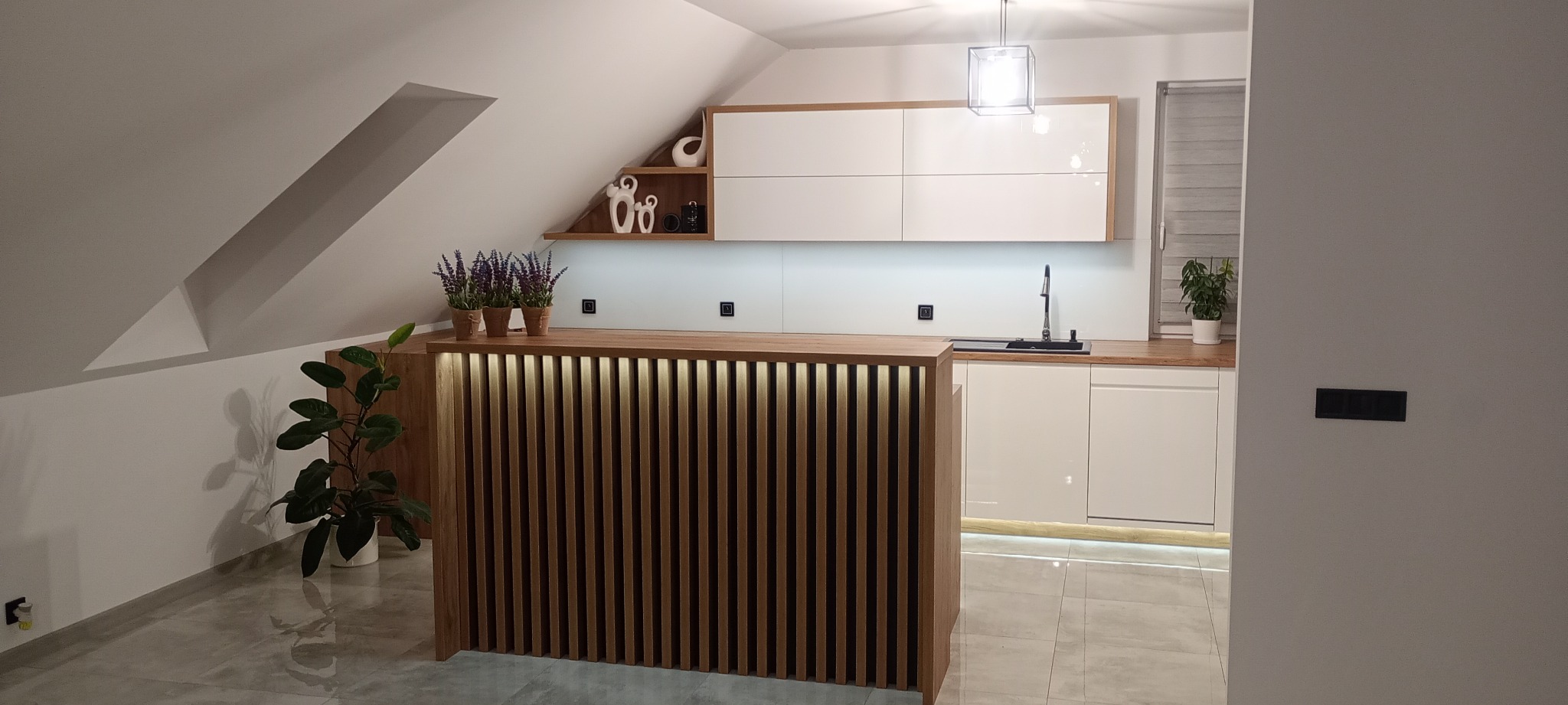 Nowoczesna kuchnia na poddaszu z białymi szafkami i drewnianym blatem. Uwagę przykuwa podświetlany, lamelowy barek i dekoracje na półce.