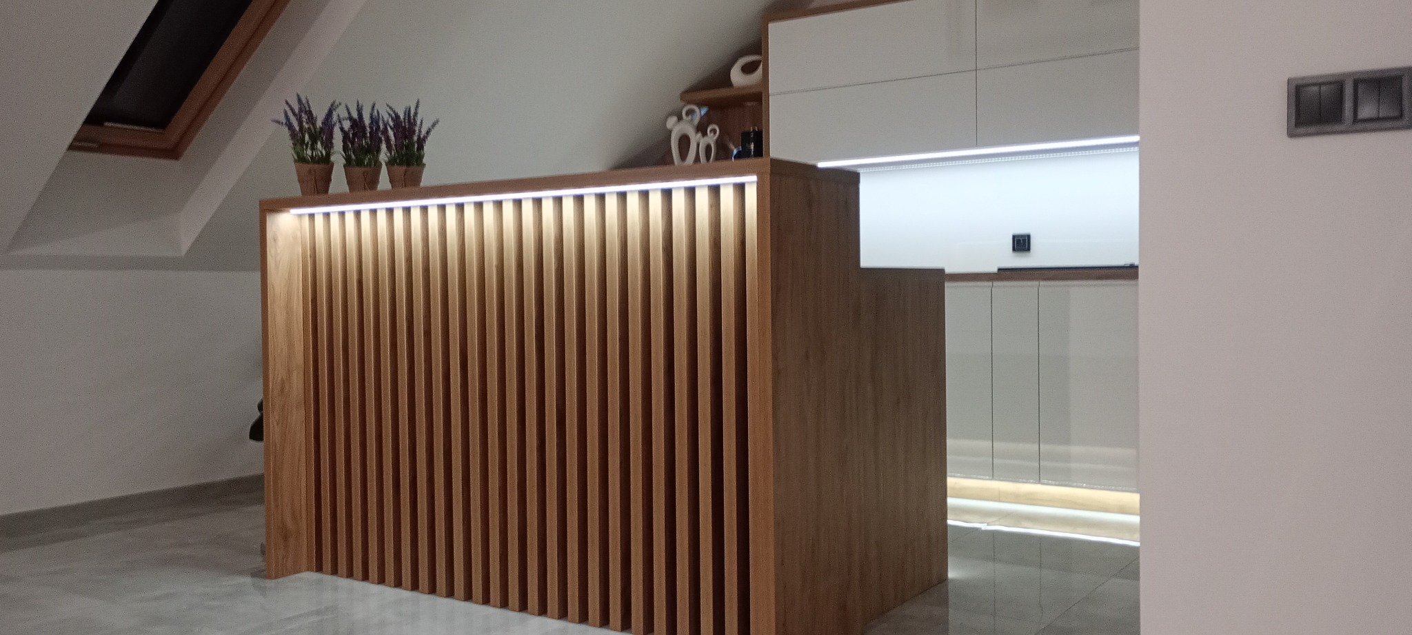 Nowoczesna zabudowa kuchenna z drewnianymi lamelami i oświetleniem LED w skosach poddasza. Na blacie doniczki z lawendą, w tle białe szafki i dekoracje.