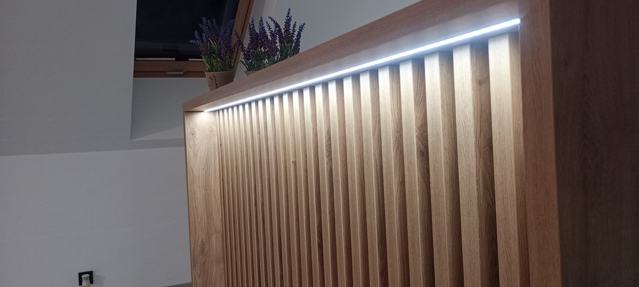 Drewniana szafa z lamelami i oświetleniem LED, zwieńczona dekoracyjnymi kwiatami w doniczkach. Ustawiona na poddaszu, widoczne okno dachowe.