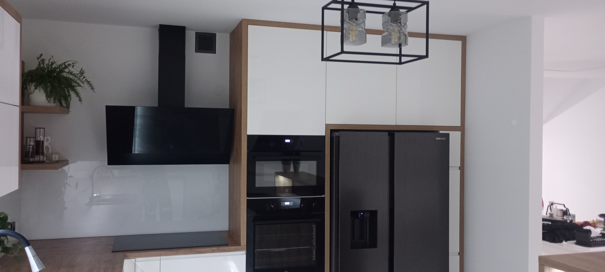 Nowoczesna kuchnia z białymi szafkami, drewnianymi akcentami i czarnym sprzętem AGD: okapem, piekarnikiem i lodówką side-by-side. Nad szafkami wisi designerska lampa.