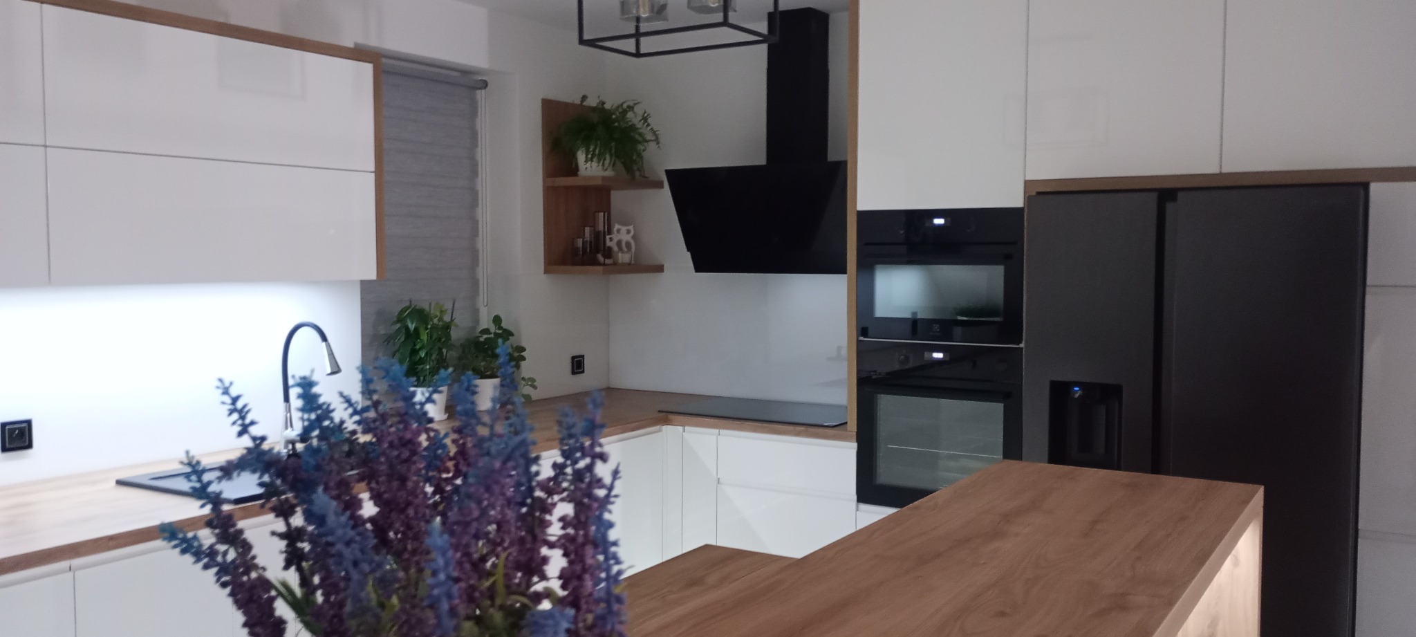 Nowoczesna kuchnia z białymi frontami, drewnianym blatem i czarnymi akcentami: okap, piekarnik, lodówka. Uwagę przyciąga bukiet kwiatów na pierwszym planie.