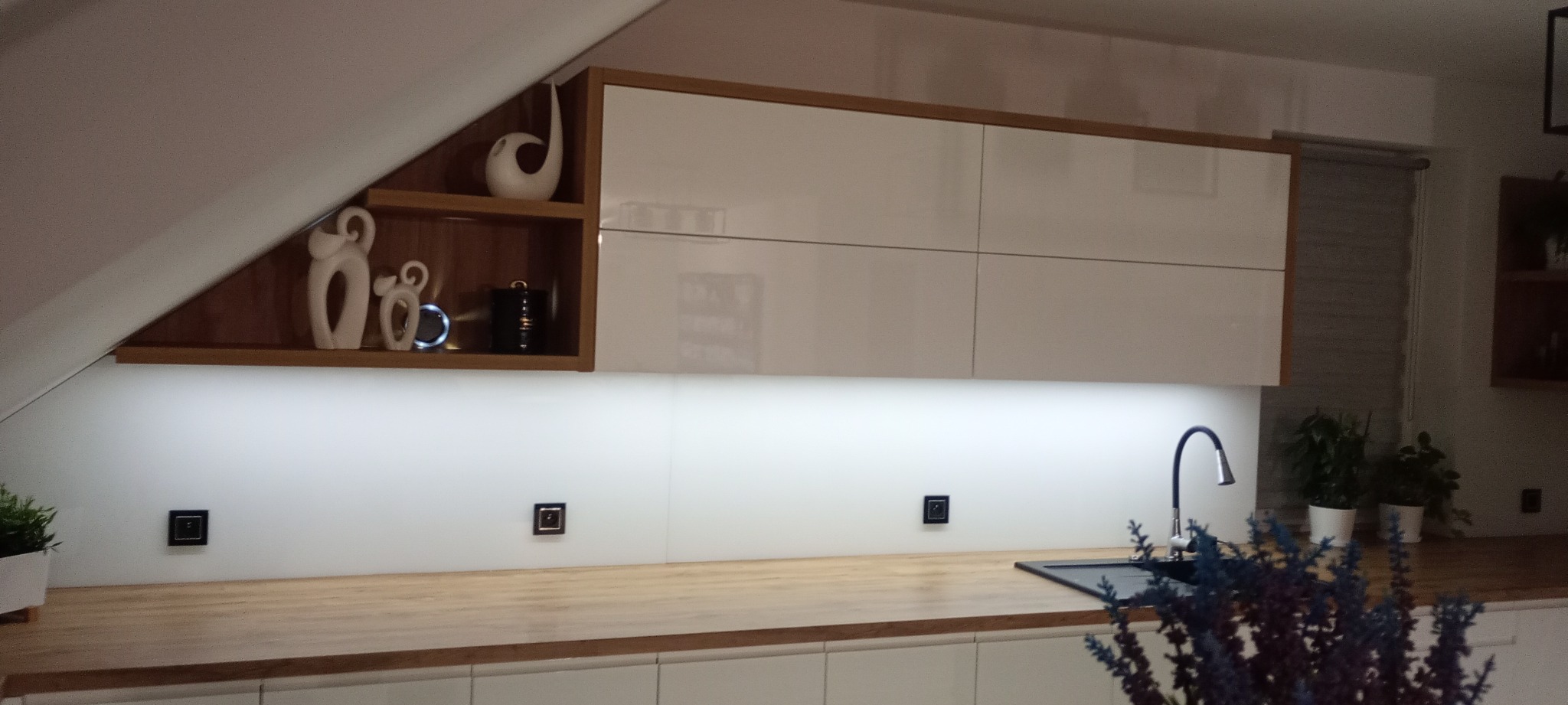 Białe szafki kuchenne pod skosem dachu z drewnianymi akcentami i oświetleniem LED. Na blacie dekoracje i kwiaty, czarna bateria.