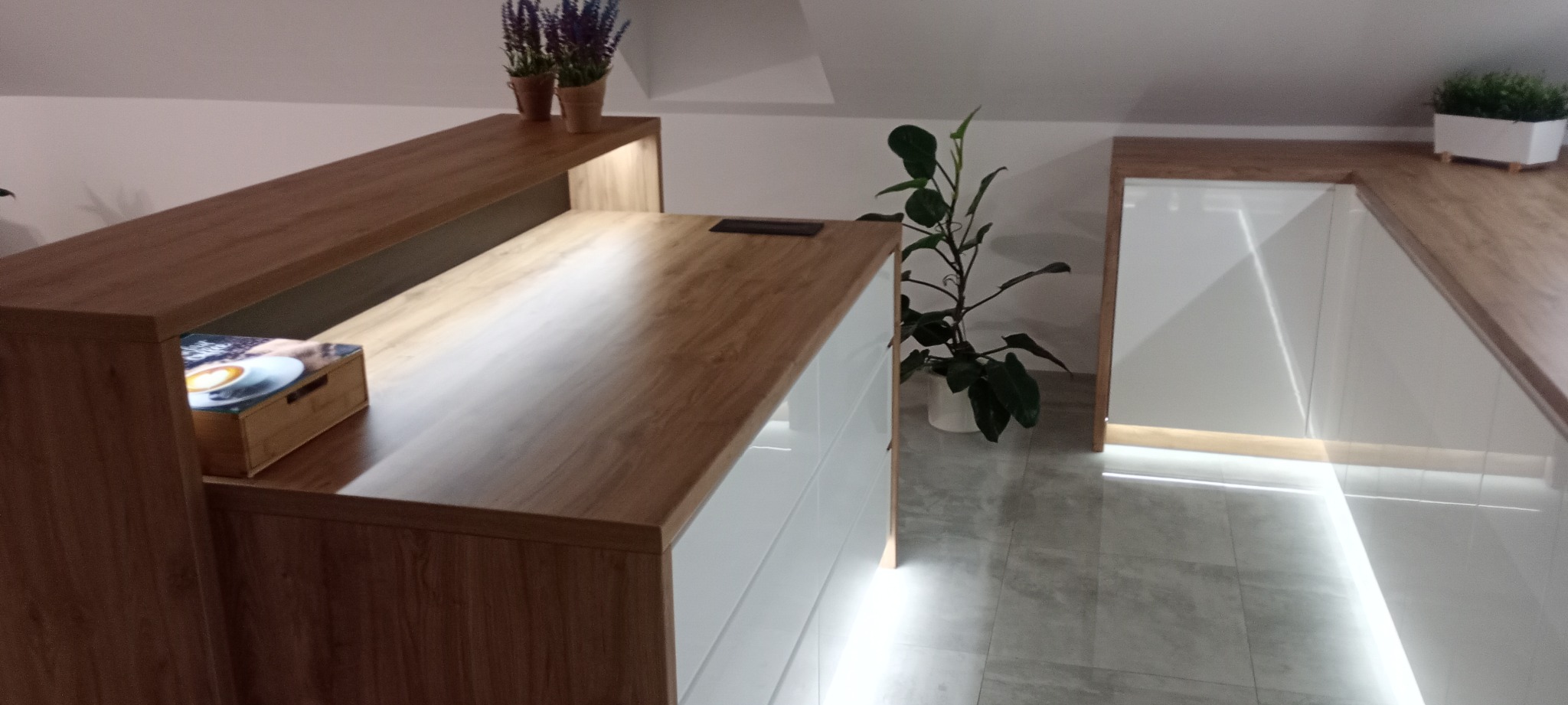 Nowoczesna zabudowa kuchenna na poddaszu z drewnianym blatem i białymi frontami. Oświetlenie LED podkreśla linię szafek. Dekoracje roślinne dodają wnętrzu świeżości.