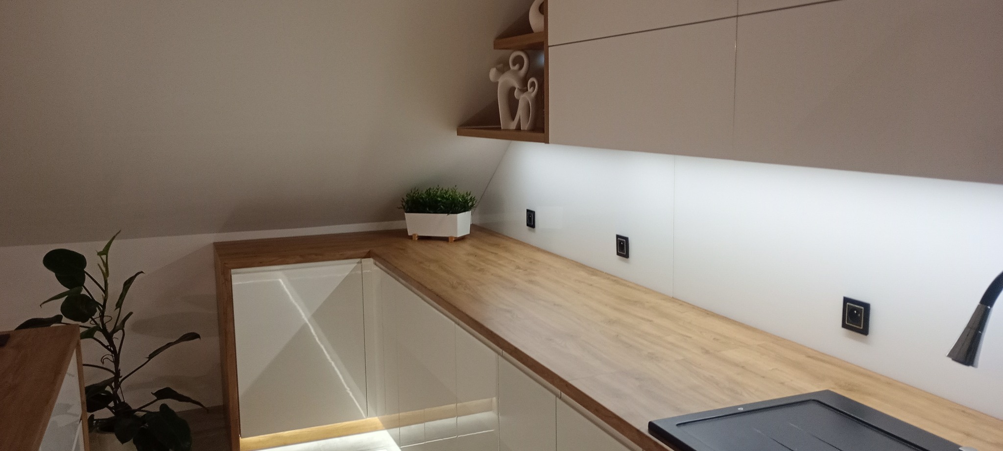 Nowoczesna kuchnia na poddaszu z białymi frontami, drewnianym blatem i oświetleniem LED. Dekoracje i roślina doniczkowa dodają przytulności.