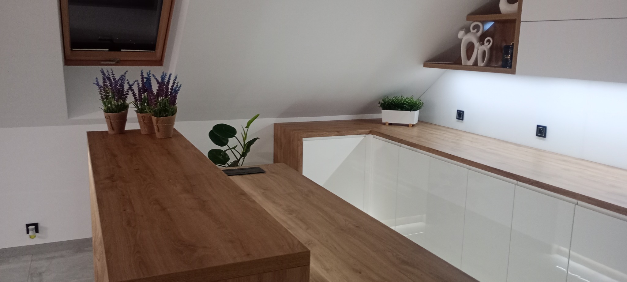 Nowoczesna zabudowa pod skosem z białymi frontami i drewnianym blatem. Uwagę przykuwają dekoracyjne rośliny doniczkowe i oświetlenie LED.