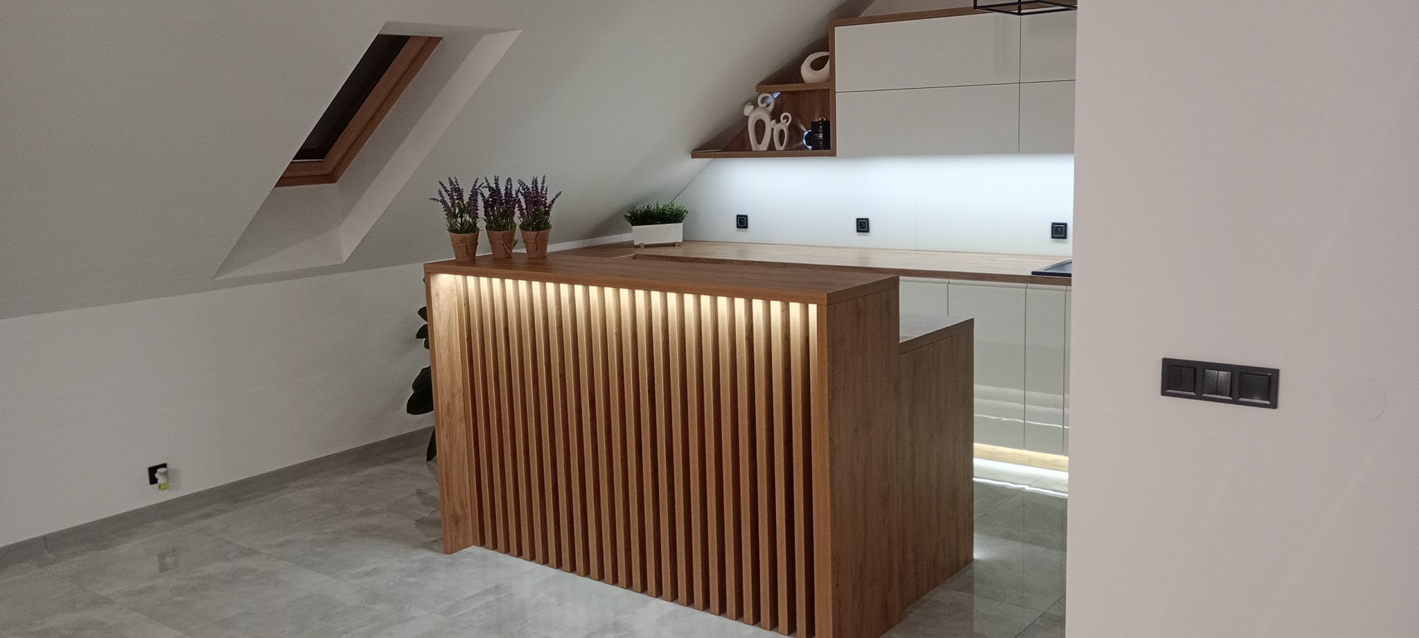 Nowoczesna kuchnia na poddaszu z drewnianym barkiem z podświetleniem LED, białe szafki i skośne okno dachowe. Minimalistyczny design.